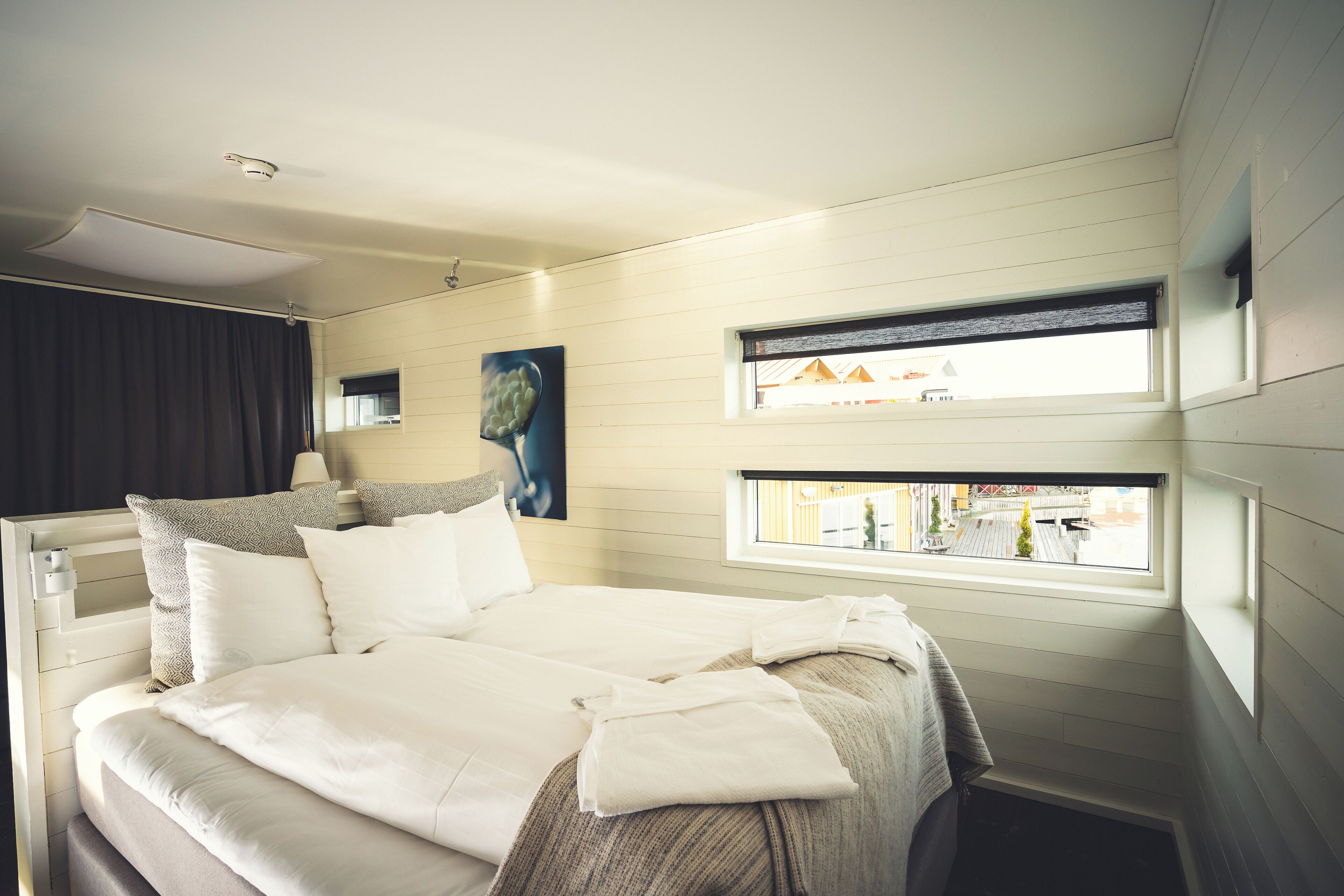 A neatly made bed with white linens, a headboard with a built-in shelf, and a window with a view of buildings outside.