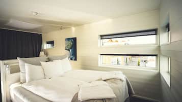 A neatly made bed with white linens, a headboard with a built-in shelf, and a window with a view of buildings outside.