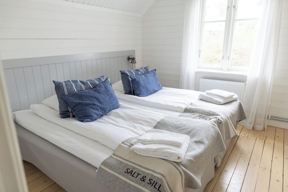 Una cama doble con dos almohadas, una mesita de noche y un ventanal con cortinas.