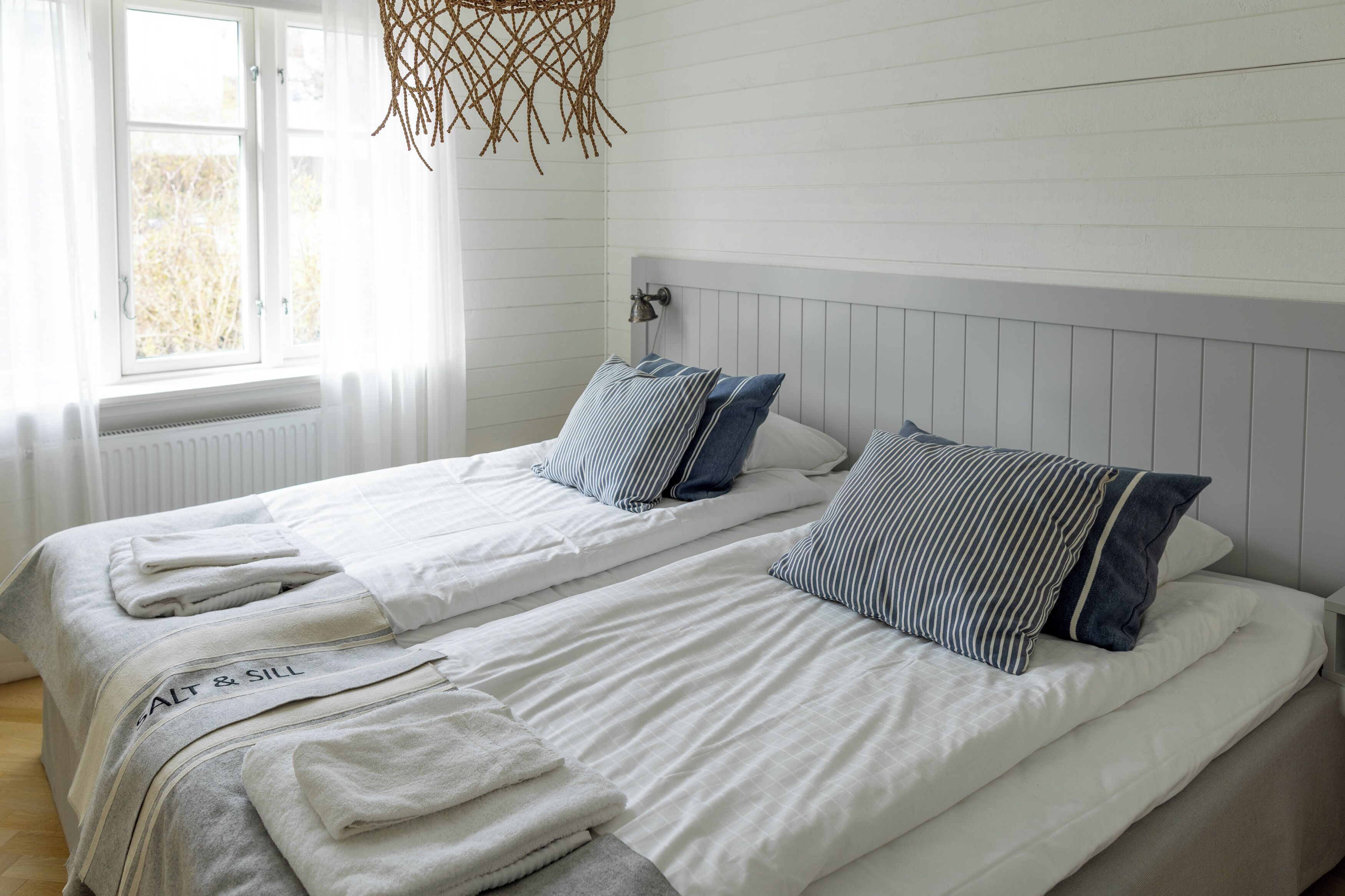 Ein Zimmer mit zwei Betten, weißer Bettwäsche, gestreifteren Kissen, einer Hängeleuchte und einem Fenster mit Jalousien.