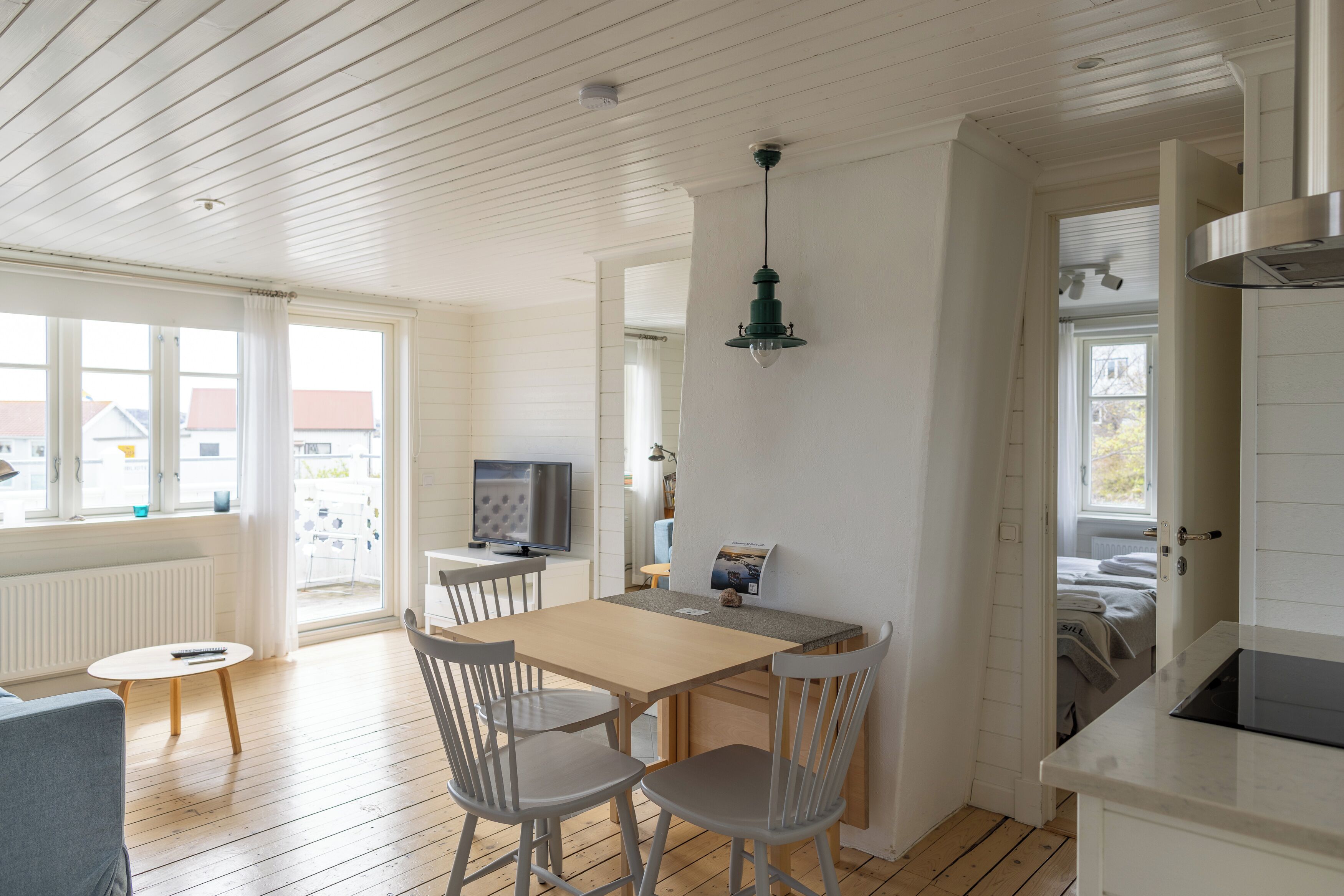 A bright kitchen with wooden floors, a dining table with chairs, a TV, and a hanging light fixture.