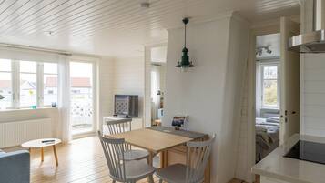 A bright kitchen with wooden floors, a dining table with chairs, a TV, and a hanging light fixture.