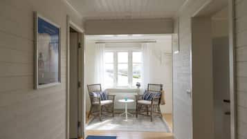 Ein Flur mit weißen Holzwänden, einem gerahmten Poster, zwei Korbstühlen mit Kissen, einem kleinen runden Tisch und einem Blick ins Freie durch ein Fenster mit weißen Vorhängen.