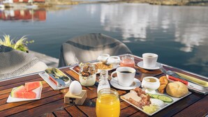 Una mesa de desayuno con diferentes alimentos, una botella de jugo de naranja y una vista a un lago tranquilo con un edificio rojo al fondo.