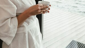 Una persona vestida de blanco sosteniendo una taza junto a una ventana con vista a un cuerpo de agua y un muelle a lo lejos.