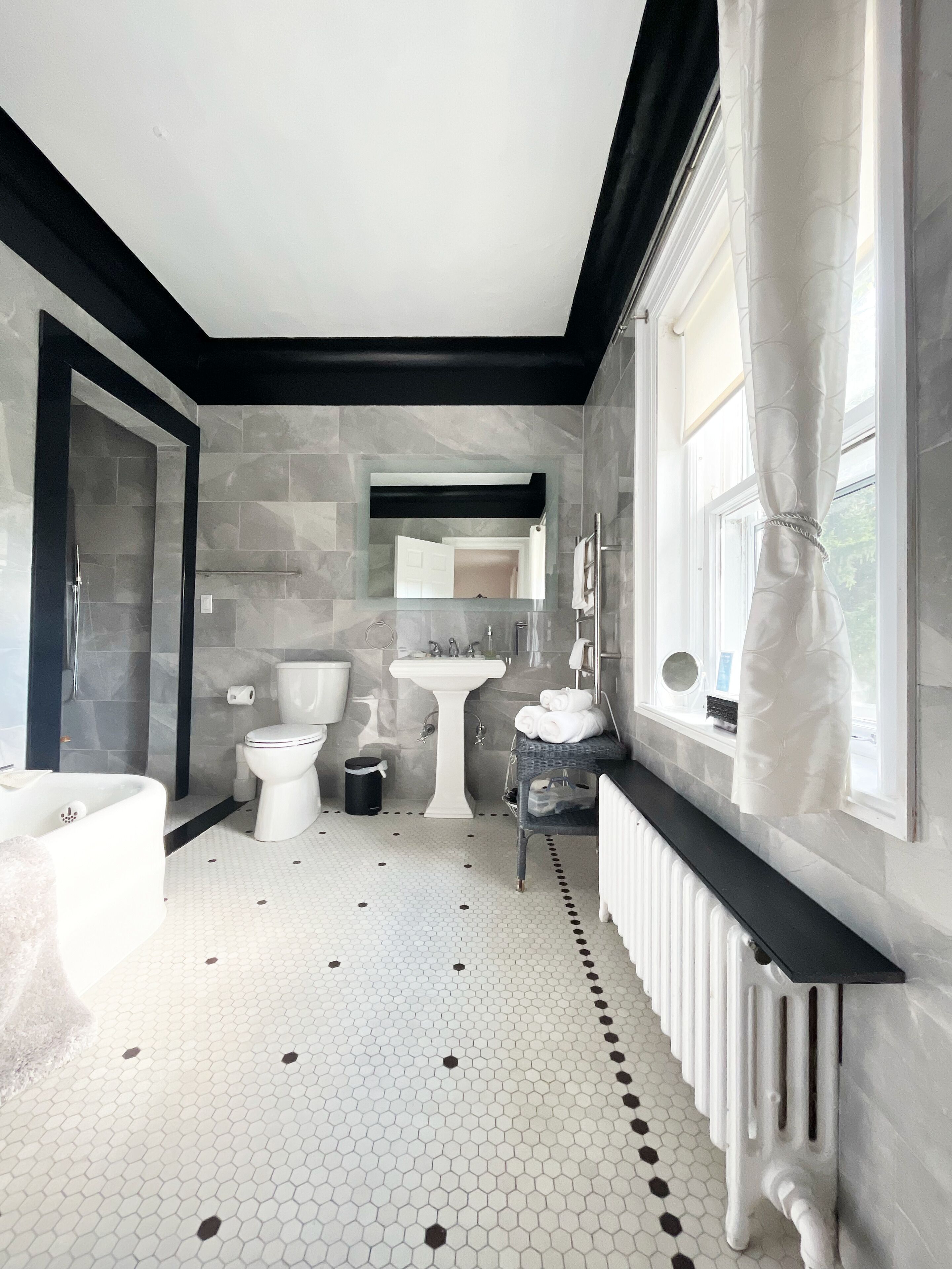 Un baño moderno con un lavamanos, inodoro y bañera blancos, un espejo con marco negro y una ventana con cortinas.