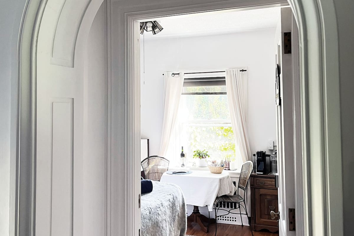 A hallway leading to a room with a bed, a table with a lamp, and a window with curtains.