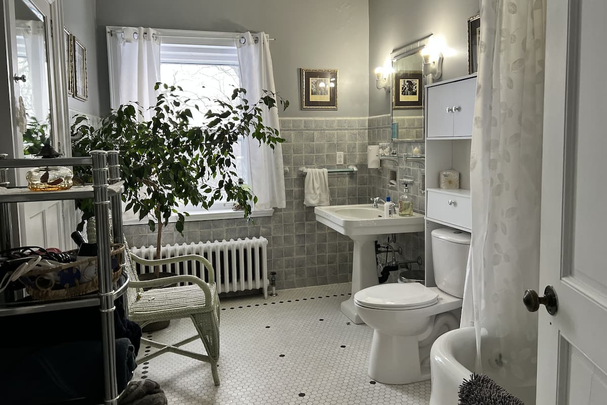 A bathroom with a white sink, toilet, and bathtub. There is a window with curtains, a radiator, and a wicker chair.