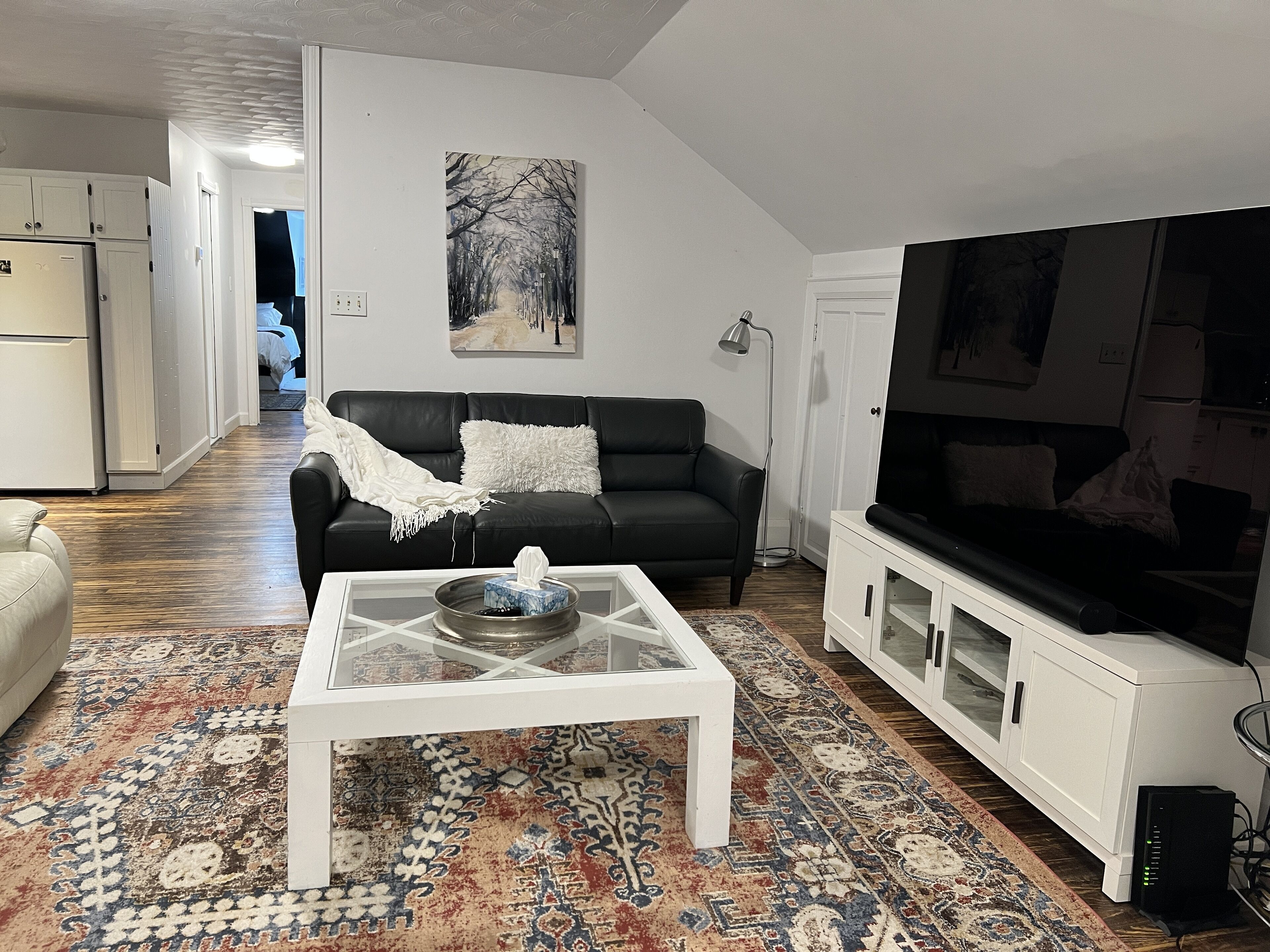 A living room with a black leather sofa, a white coffee table, a large flat-screen TV, and a patterned rug.