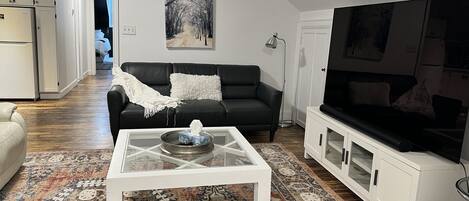A living room with a black leather sofa, a white coffee table, a large flat-screen TV, and a patterned rug.