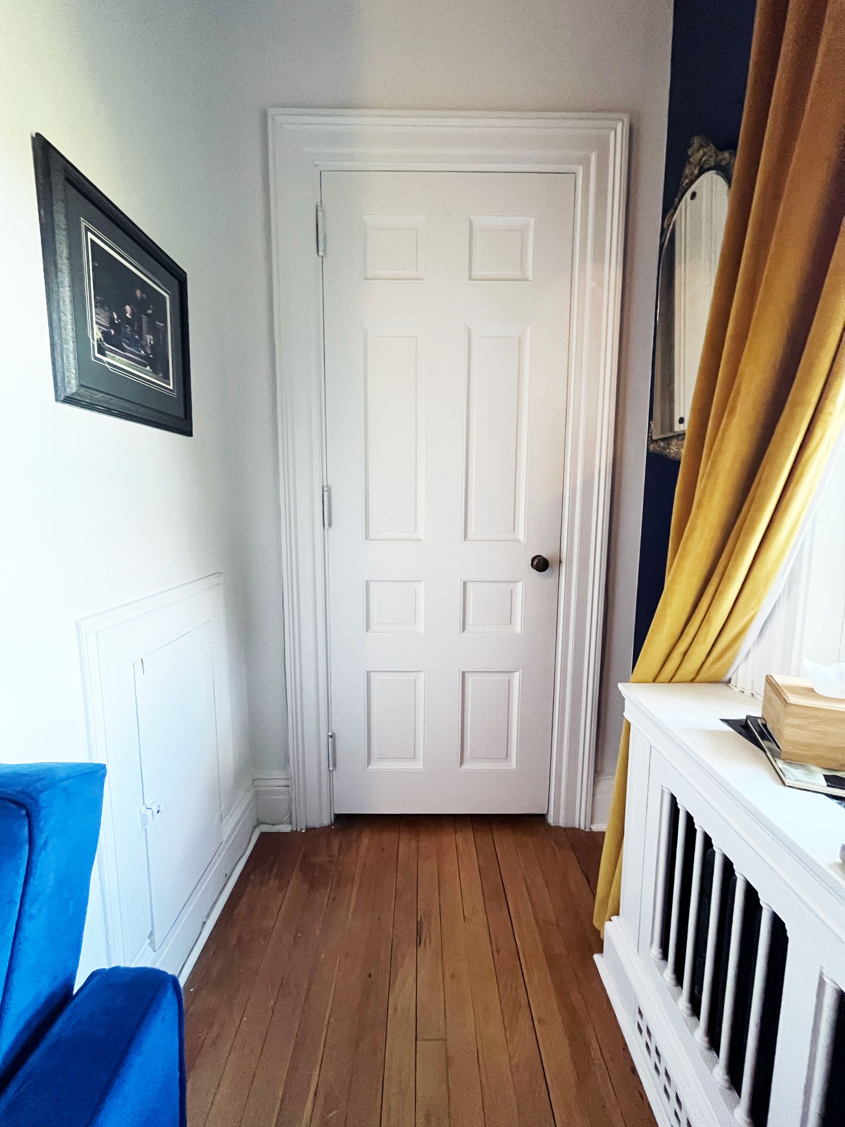 Un couloir avec une porte blanche, une chaise bleue, un rideau jaune et une cheminée blanche.