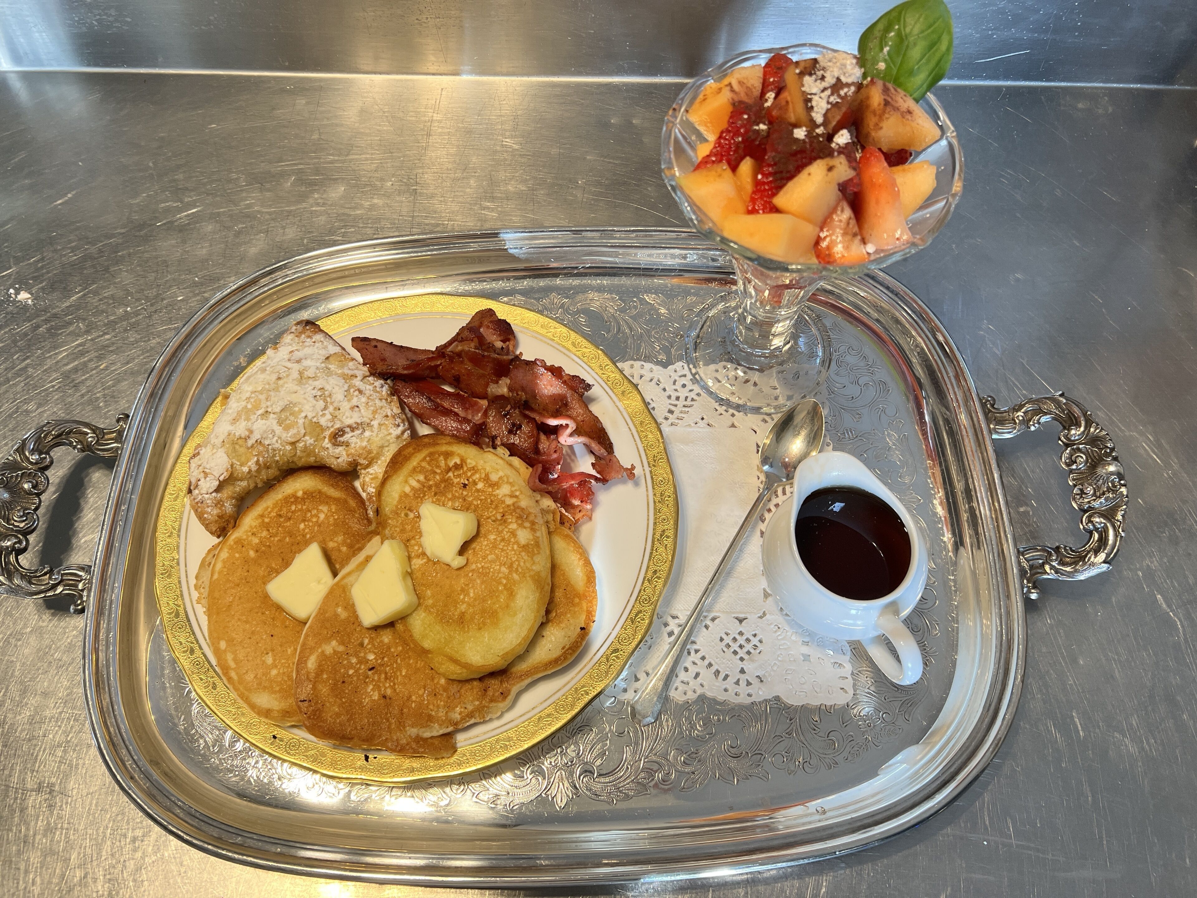 Un plateau de petit-déjeuner avec des crêpes, du bacon et une salade de fruits, posé sur une table en acier inoxydable.