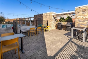 Un patio exterior con mesas, sillas, una parrilla y luces colgantes.