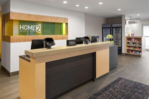 A hotel reception desk with a digital kiosk, a minibar, and a refrigerator stocked with snacks. - Home2 Suites by Hilton Meridian (Meridian)