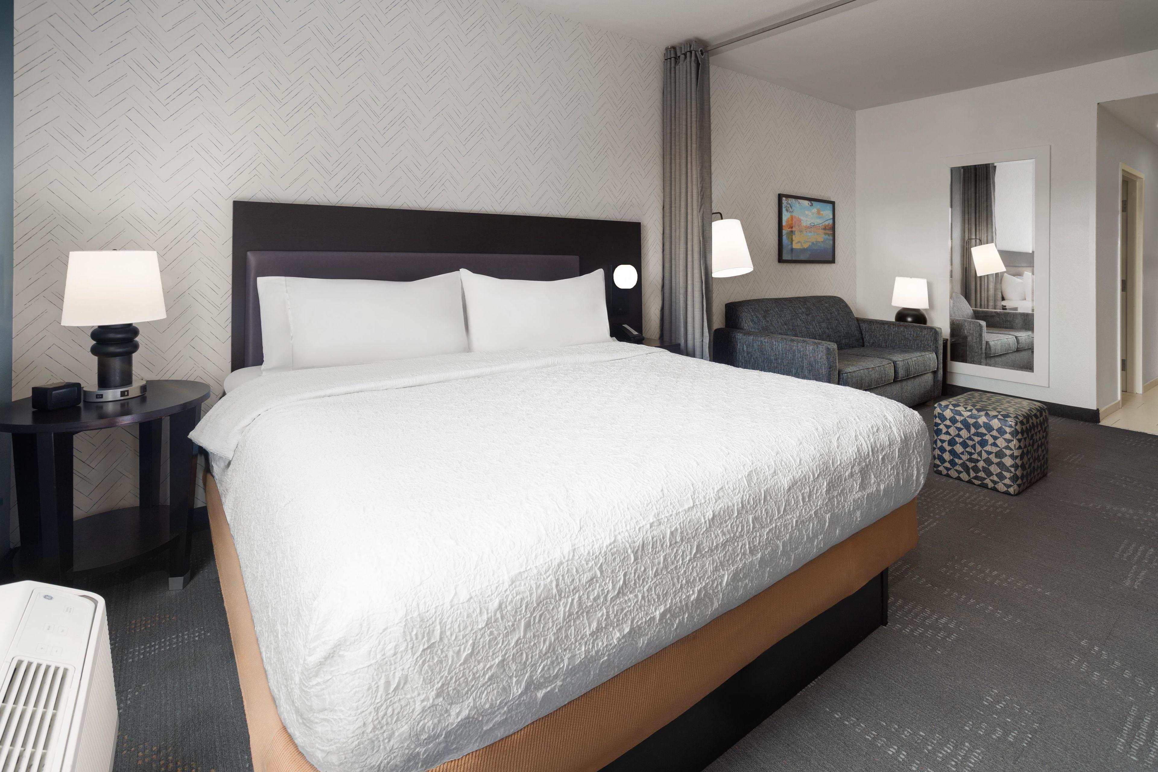 A hotel room with a large bed, two bedside tables with lamps, a grey sofa, a patterned ottoman, and a mirror.