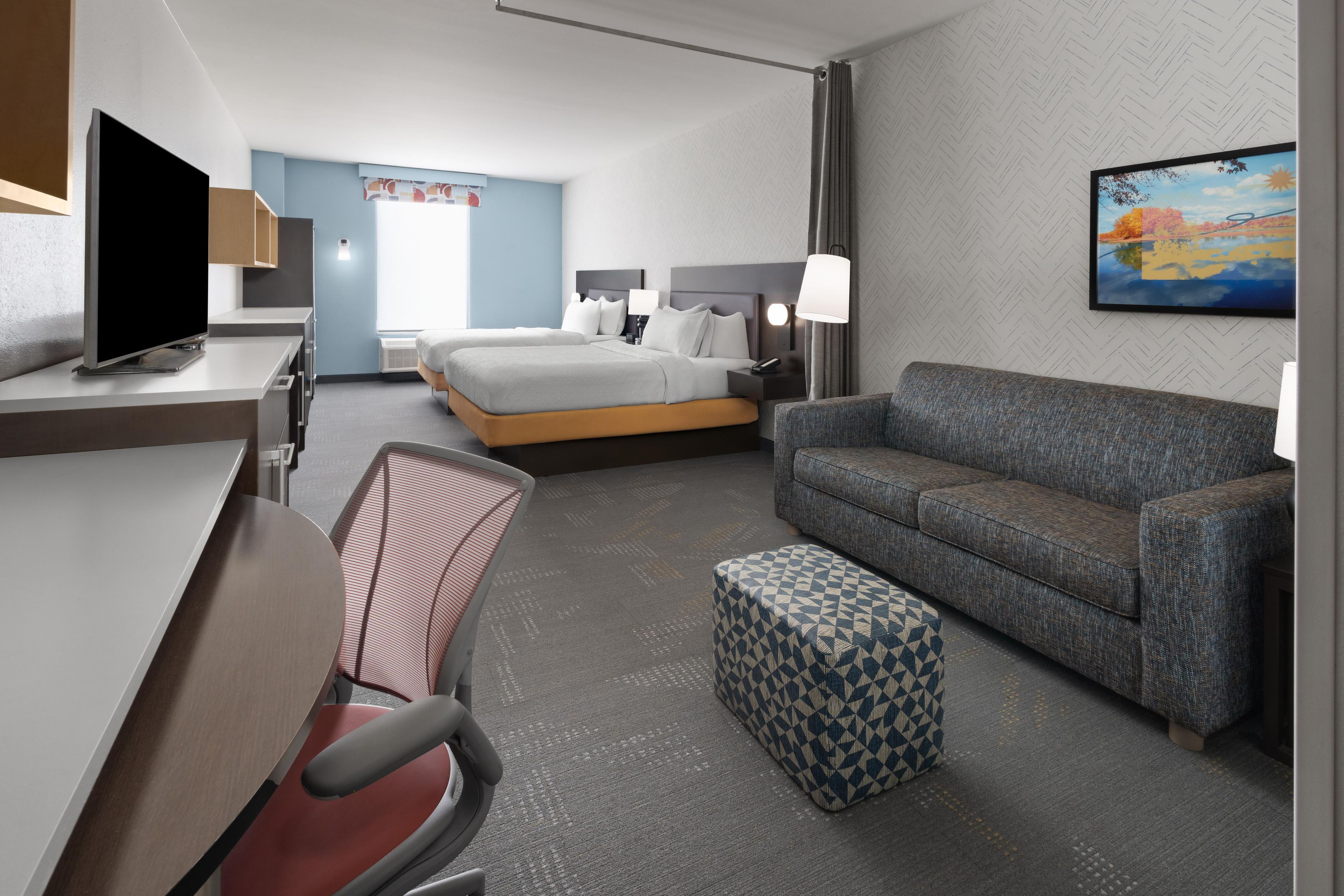 A hotel room with a bed, a sofa, a desk with a TV, a chair, and a small ottoman.