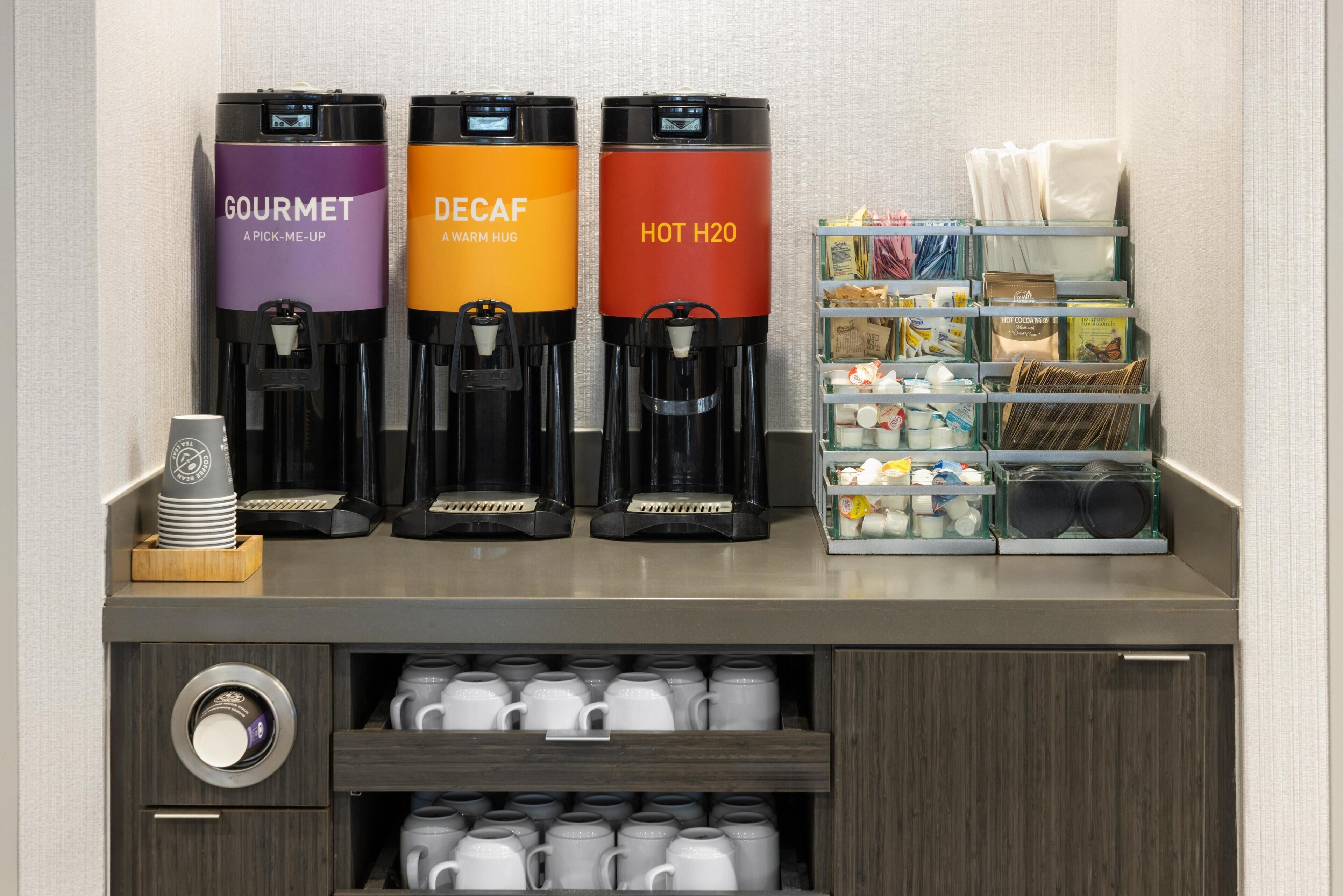 Een koffiehoek met drie dispensers, gelabeld 'GOURMET', 'DECAF' en 'HEET WATER'.