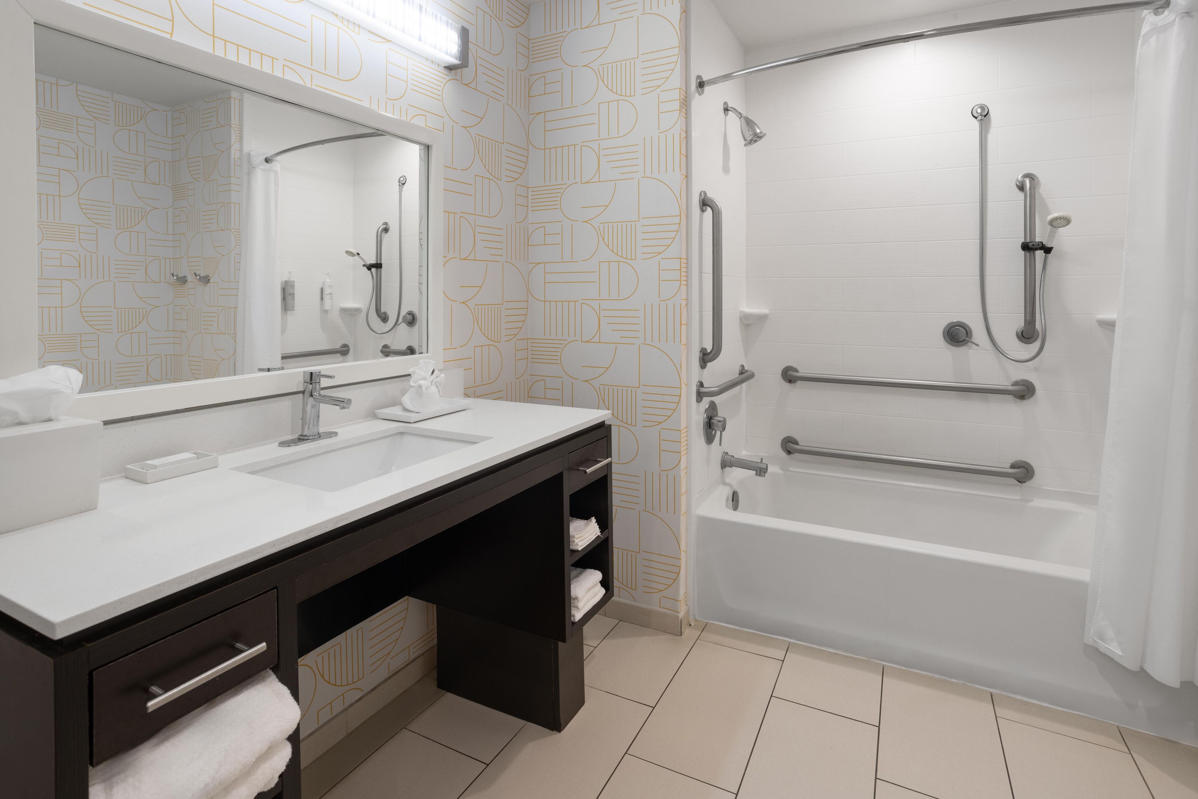 A modern bathroom with a double sink vanity, a large mirror, a shower with grab bars, and a bathtub.