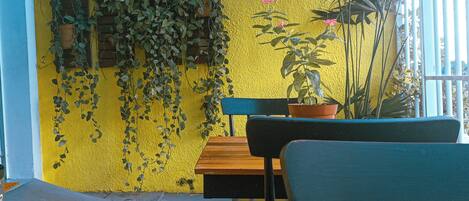 A yellow wall with potted plants, a wooden table, and chairs in a covered outdoor area.