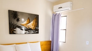 Une chambre d’hôtel avec un lit, du linge de lit blanc, une tête de lit marron, un tableau représentant un voilier, un appareil de climatisation et une fenêtre avec des rideaux violets.