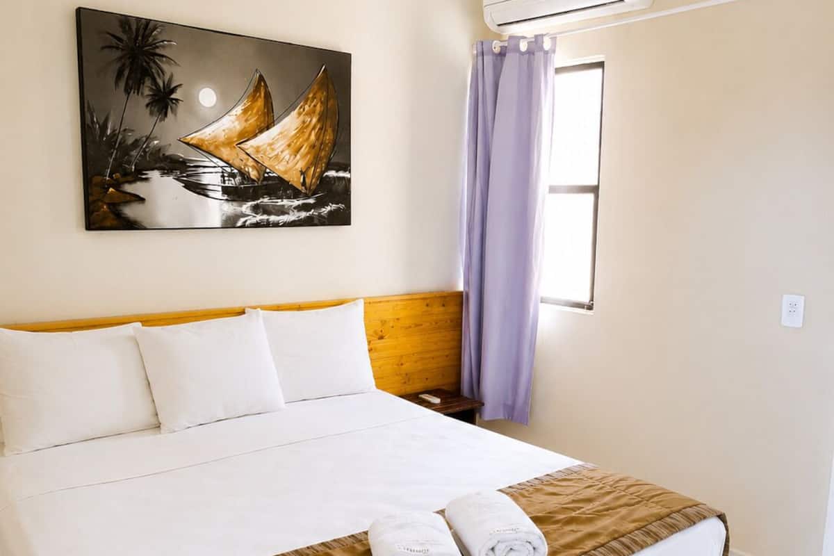 Habitación de hotel con cama, ropa de cama blanca, cabecera marrón, un cuadro de un velero, unidad de aire acondicionado y una ventana con cortinas moradas.