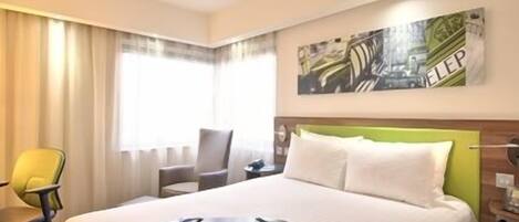Una habitación de hotel con cama, escritorio, silla y ventana con cortinas.
