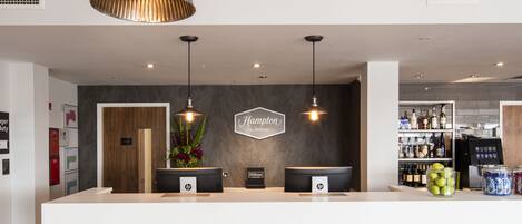 A hotel reception area with a modern design, featuring a reception desk, a sign that reads "Hampton by Hilton," and a bar area with various bottles and a refrigerator.