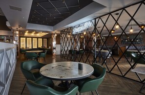 A restaurant interior with a modern design, featuring a mix of round and rectangular tables, green upholstered chairs, and a brick accent wall. - Hampton by Hilton Humberside Airport (Kirmington)