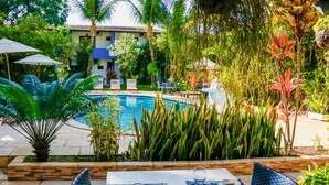Un área de comedor junto a la piscina con mesas dispuestas para dos, rodeada de exuberante vegetación tropical y una vista de un edificio al fondo.