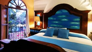 Un dormitorio con una cama grande, una mesita de noche con lámpara, un cabecero de madera con un cuadro del océano y vistas de un jardín a través de una ventana.