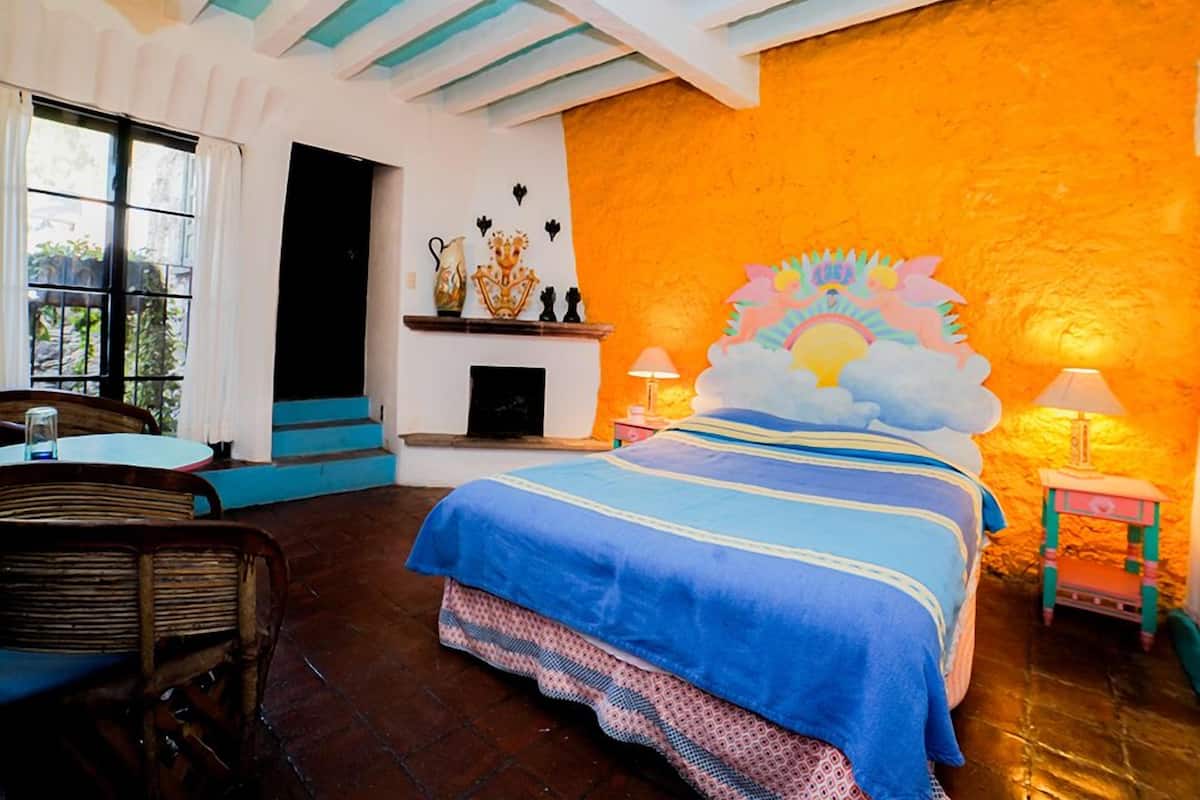 A room with a bed, a table, chairs, a fireplace, and a mural on the wall.