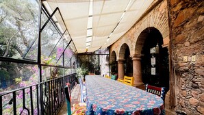 Un área de comedor al aire libre con techo, con una mesa dispuesta para dos personas, rodeada de muros y arcos de piedra, y vistas a árboles y flores a través de amplios ventanales.