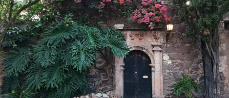 A stone building with a black door, surrounded by greenery and a stone wall.