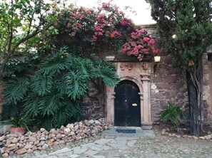 A stone building with a black door, surrounded by greenery and a stone wall.