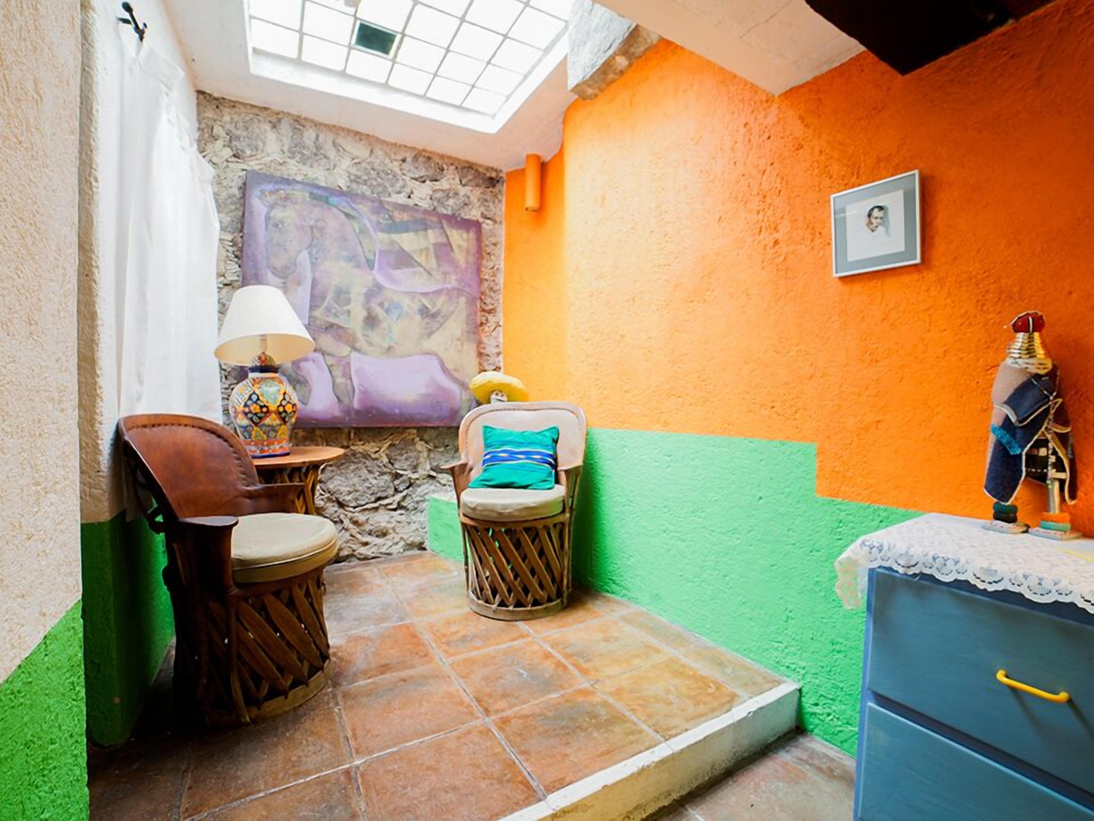 A cozy room with a stone wall, a colorful painting, a small table with a lamp, a chair, a wooden cabinet, and a small statue.