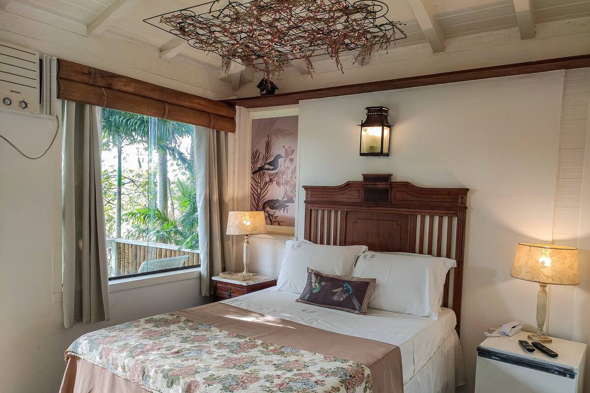 Una habitación de hotel con una cama, una mesita de noche, una lámpara, luz en la pared, una ventana con cortinas e iluminación en el techo.
