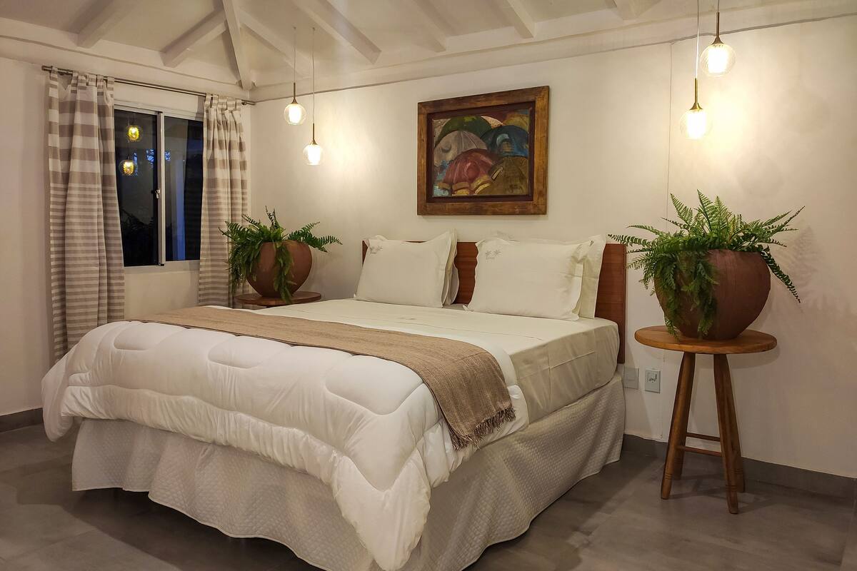 Un dormitorio con una cama grande, dos plantas en macetas, un cuadro en la pared y una ventana con cortinas.