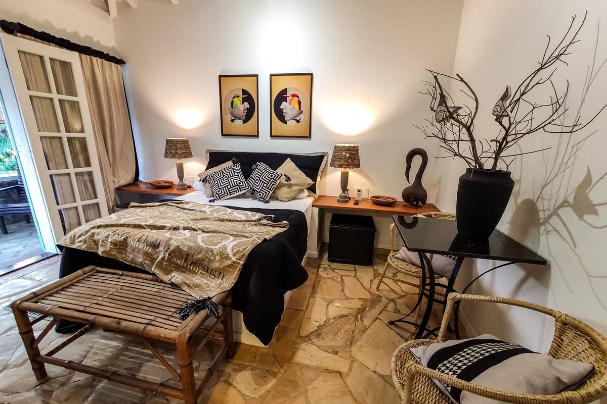 Un dormitorio con cama, mesitas de noche, escritorio, silla, lámpara, un jarrón con ramas y dos cuadros decorativos.