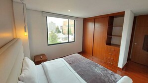 A modern bedroom with a large bed, a wardrobe, and a window with a city view. - Luxury Suites & Apartments (Bogotá)