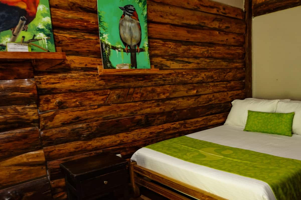A wooden cabin-style room with a bed, a nightstand, and a wall-mounted painting of a bird.
