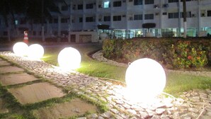 A pathway with illuminated spherical lights, a building with multiple windows, and a landscaped area with grass and plants.