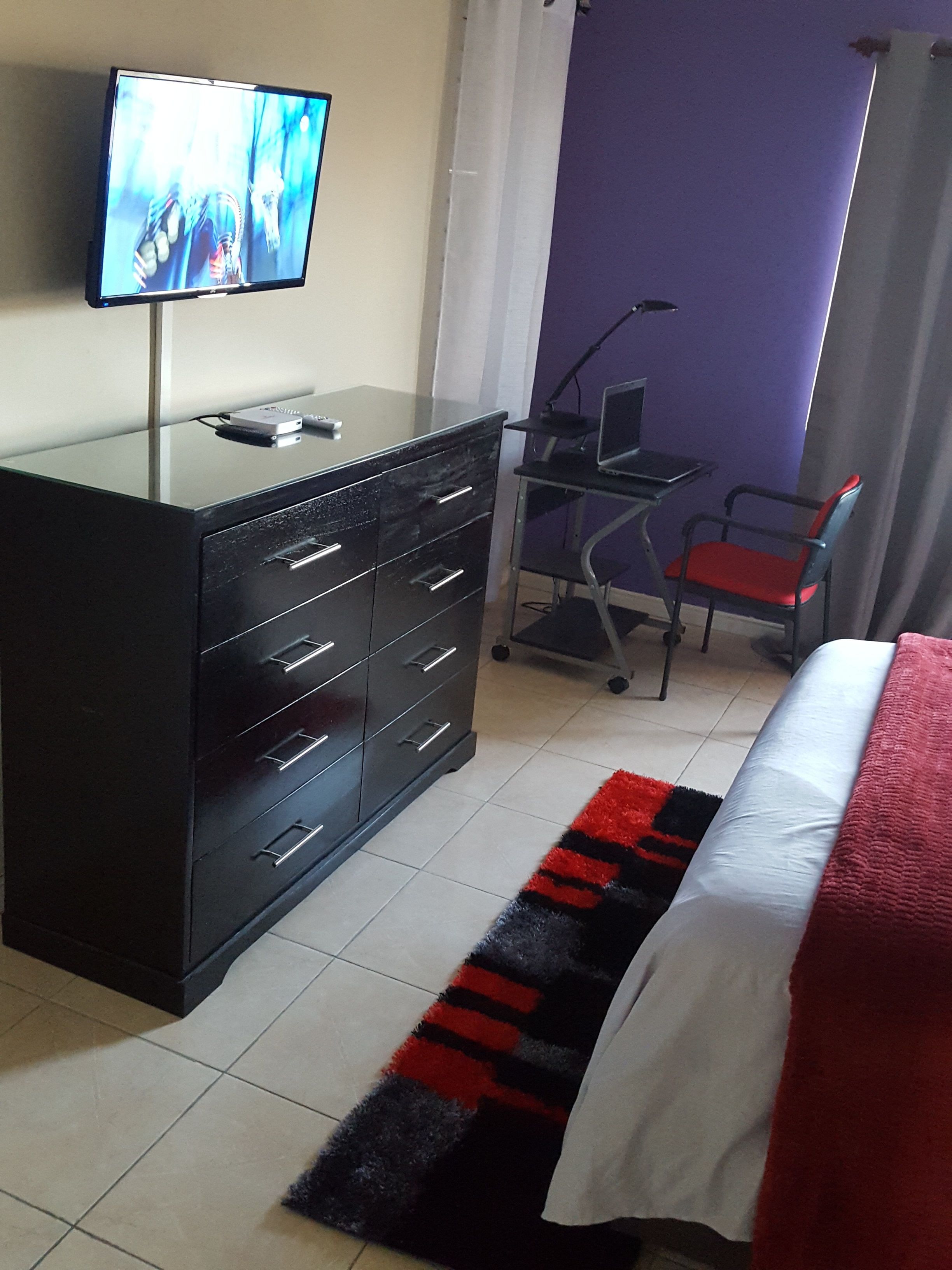 A hotel room with a bed, a TV on a dresser, a desk with a chair, and a small rug.