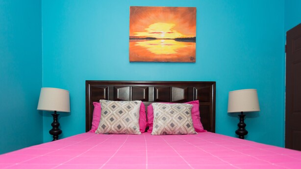 Een kamer met een helderblauwe wand, een roze sprei en een zonsondergangschilderij boven het bed.