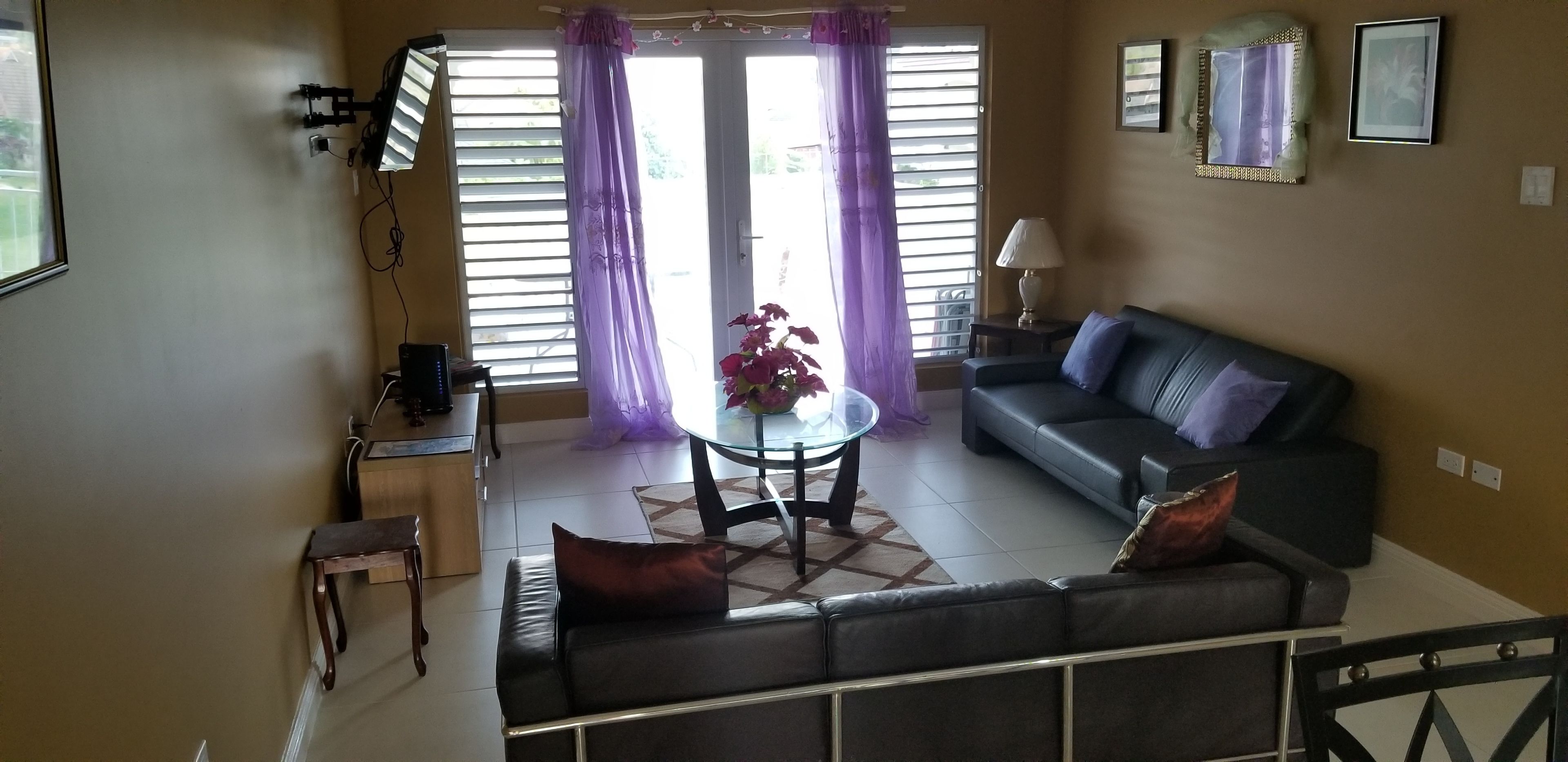 A living room with a sofa, a glass coffee table, a small table with a lamp, and a window with purple curtains.