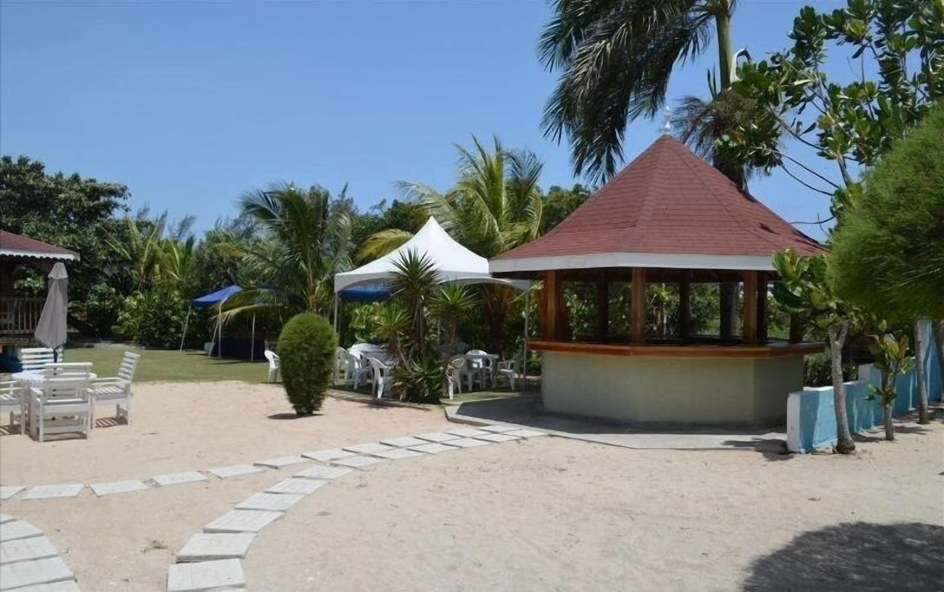 Un pabellón con techo de tejas, rodeado de palmeras y una zona arenosa con sillones blancos.