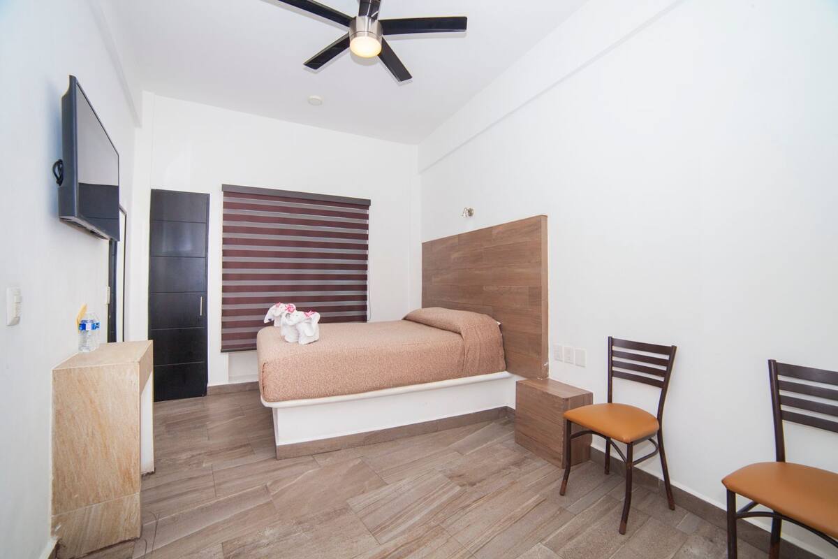 Quarto com cama, televisão, ventilador de teto e janela com persianas.
