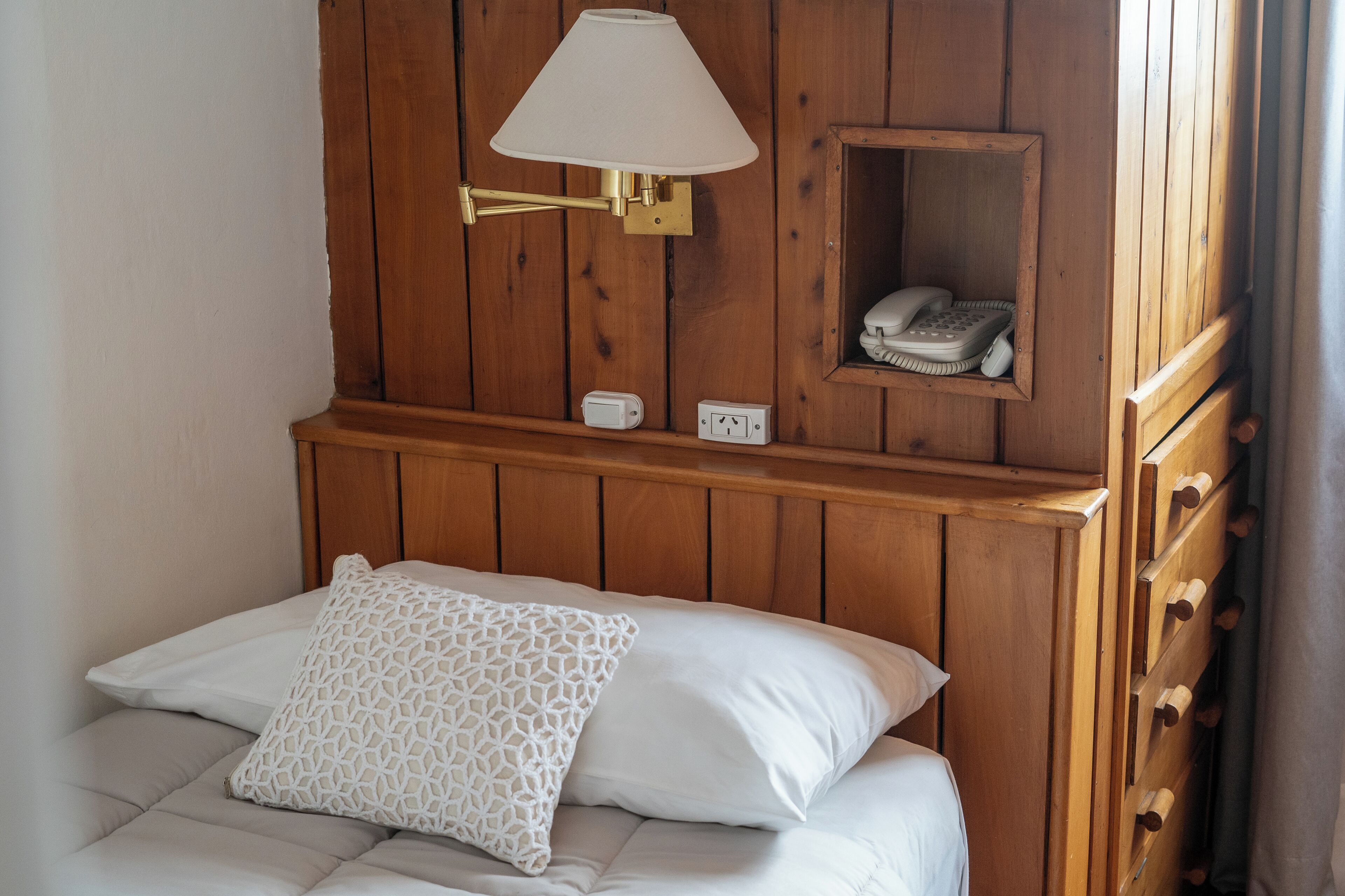 A bed with white linens and a patterned pillow, a wall-mounted lamp, a telephone, and a power outlet.