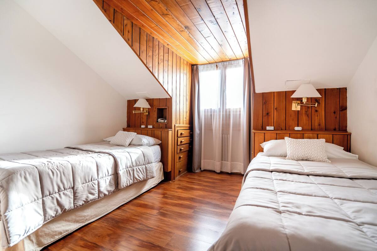 A room with two beds, wooden paneling on the walls, a ceiling fan, and a window with curtains.