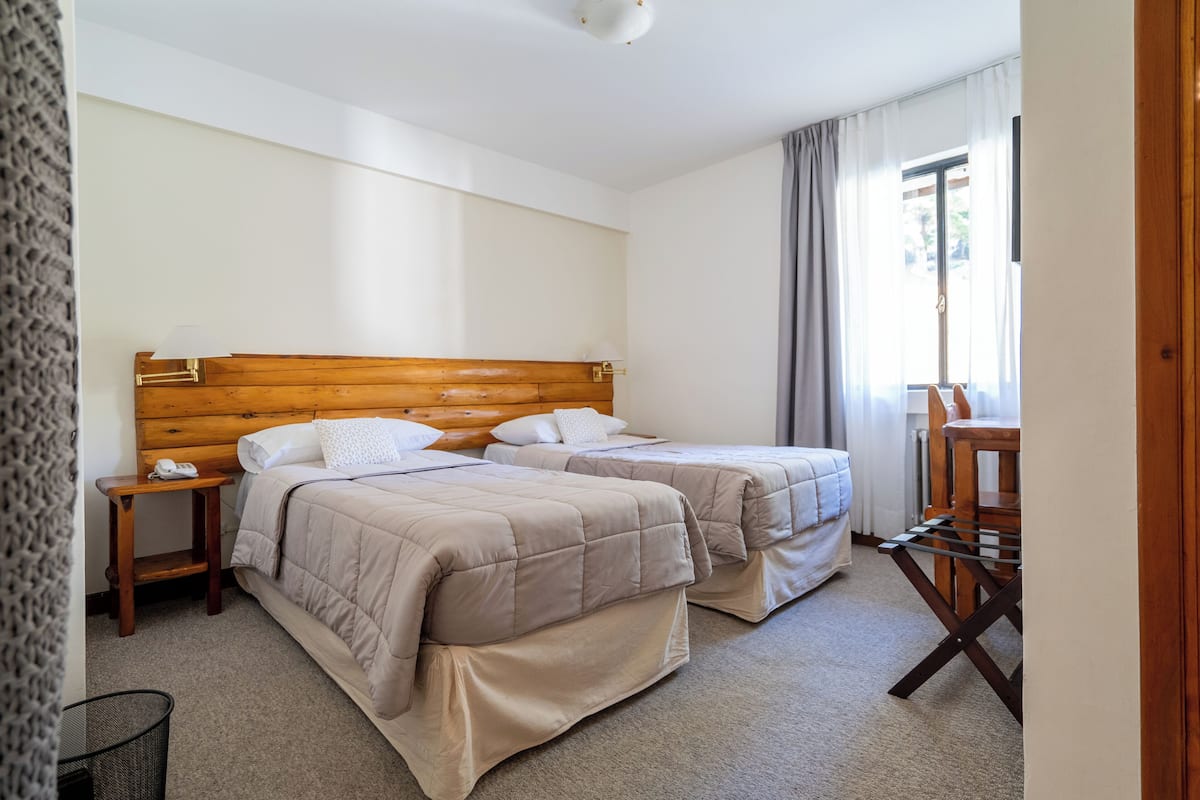 A bedroom with two beds, a wooden headboard, a bedside table with a lamp, and a window with curtains.