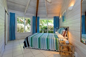 A bedroom with a bed, a nightstand, a mirror, and a window with curtains. - Sands Villas Rarotonga (Rarotonga)